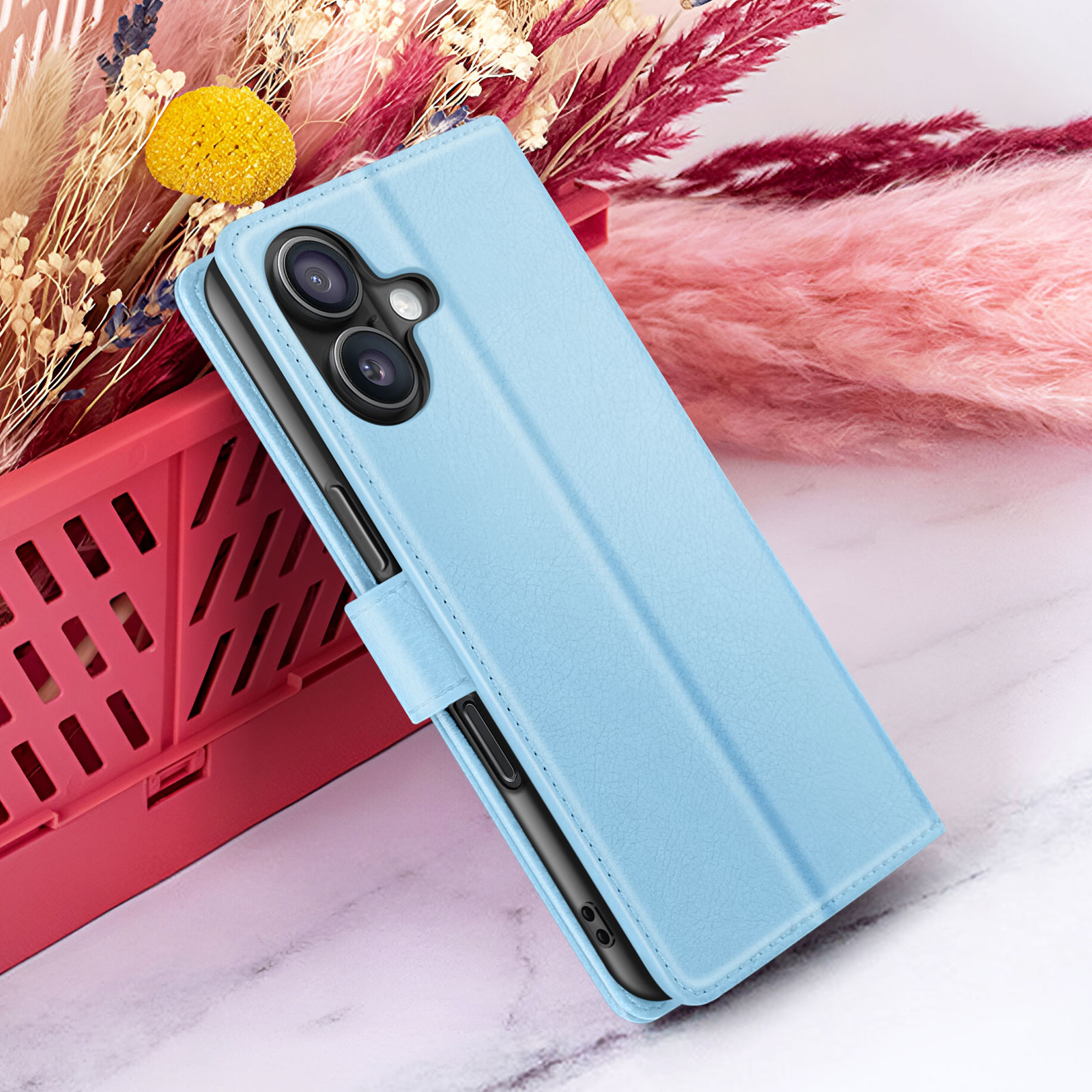 Een lichtblauwe telefoonhoes is open, met een telefoon met cameralenzen. Achtergrond inclusief bloemen en een rode container.