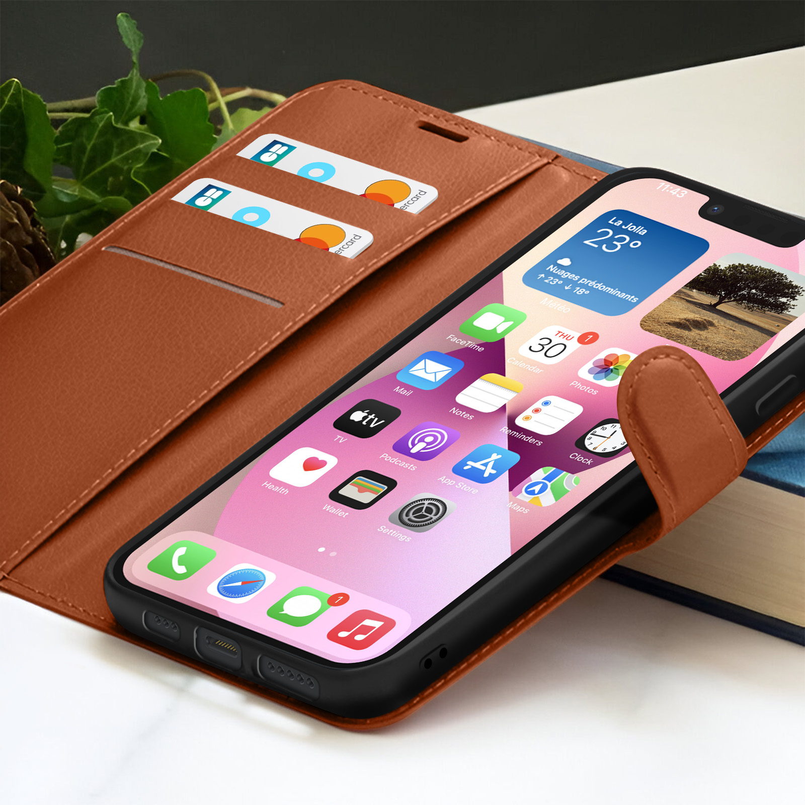 Een iPhone in een bruine leren hoes toont apps. Creditcards in een zak. Boek en bladeren op de achtergrond.