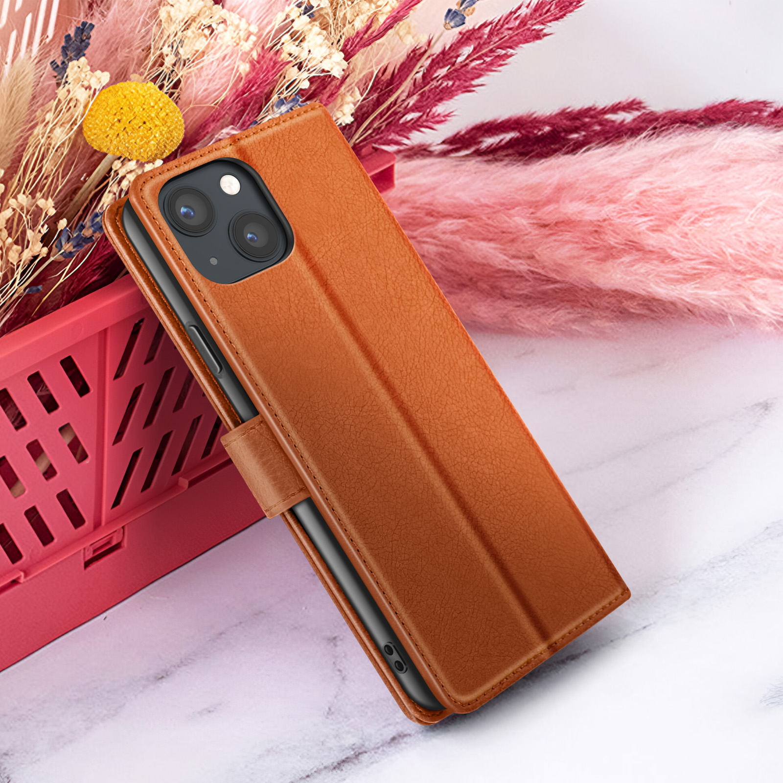 Bruine leren telefoonhoes met telefoon erin, leunend tegen bloemen. Achtergrond bevat roze en witte elementen.