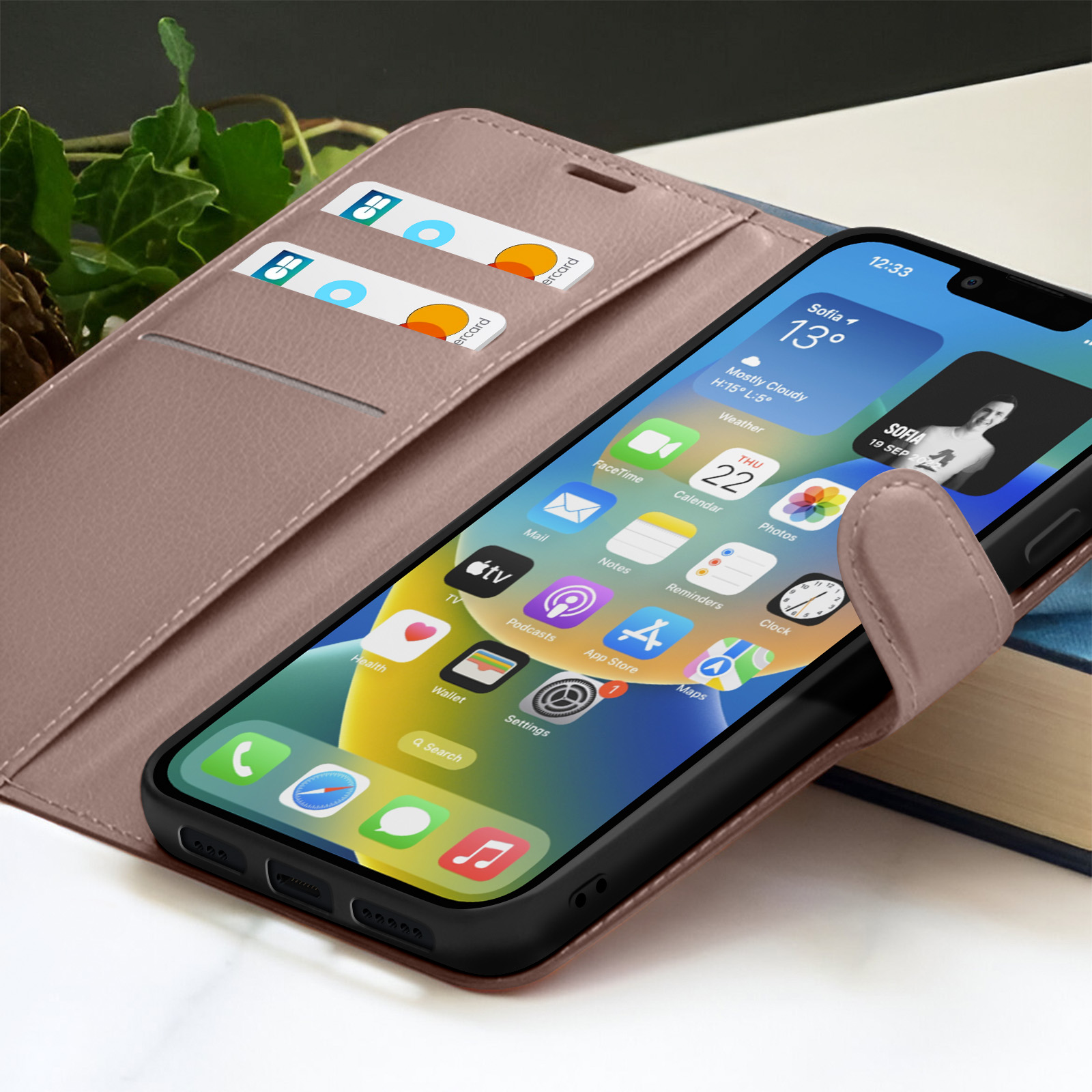 Een iPhone in een bruine hoes toont apps. Twee kaarten zitten in de hoes. Achtergrond heeft bladeren en een boek.
