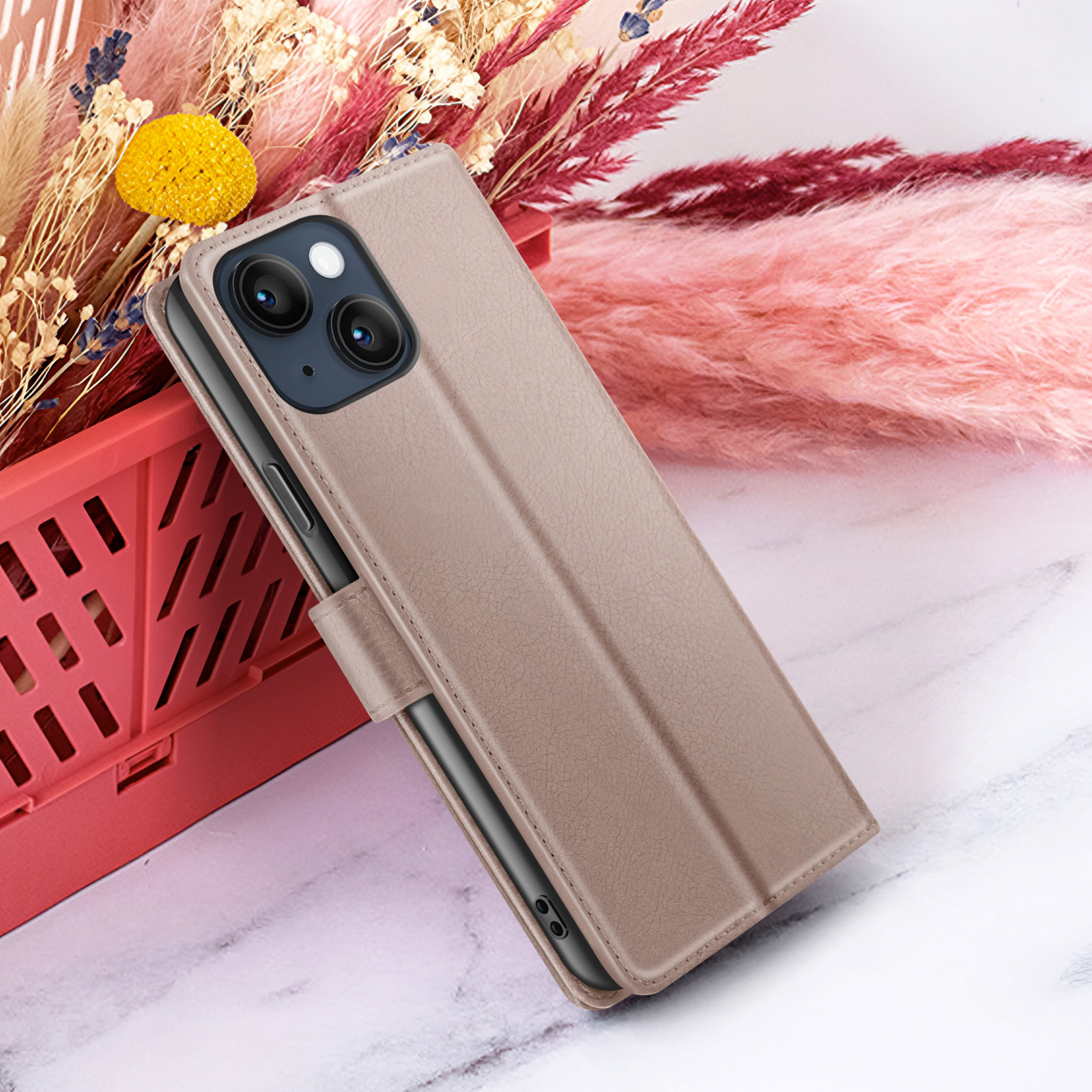 Een telefoonhoesje van beige leer wordt getoond. Een bloemstuk op de achtergrond bevat roze, gele en bordeauxrode kleuren.