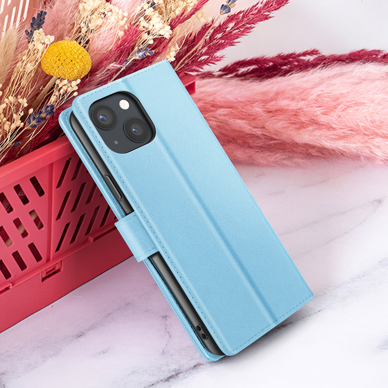 Un étui de téléphone bleu clair avec un téléphone à l'intérieur est sur une surface en marbre, près de fleurs séchées et d'un conteneur rouge.