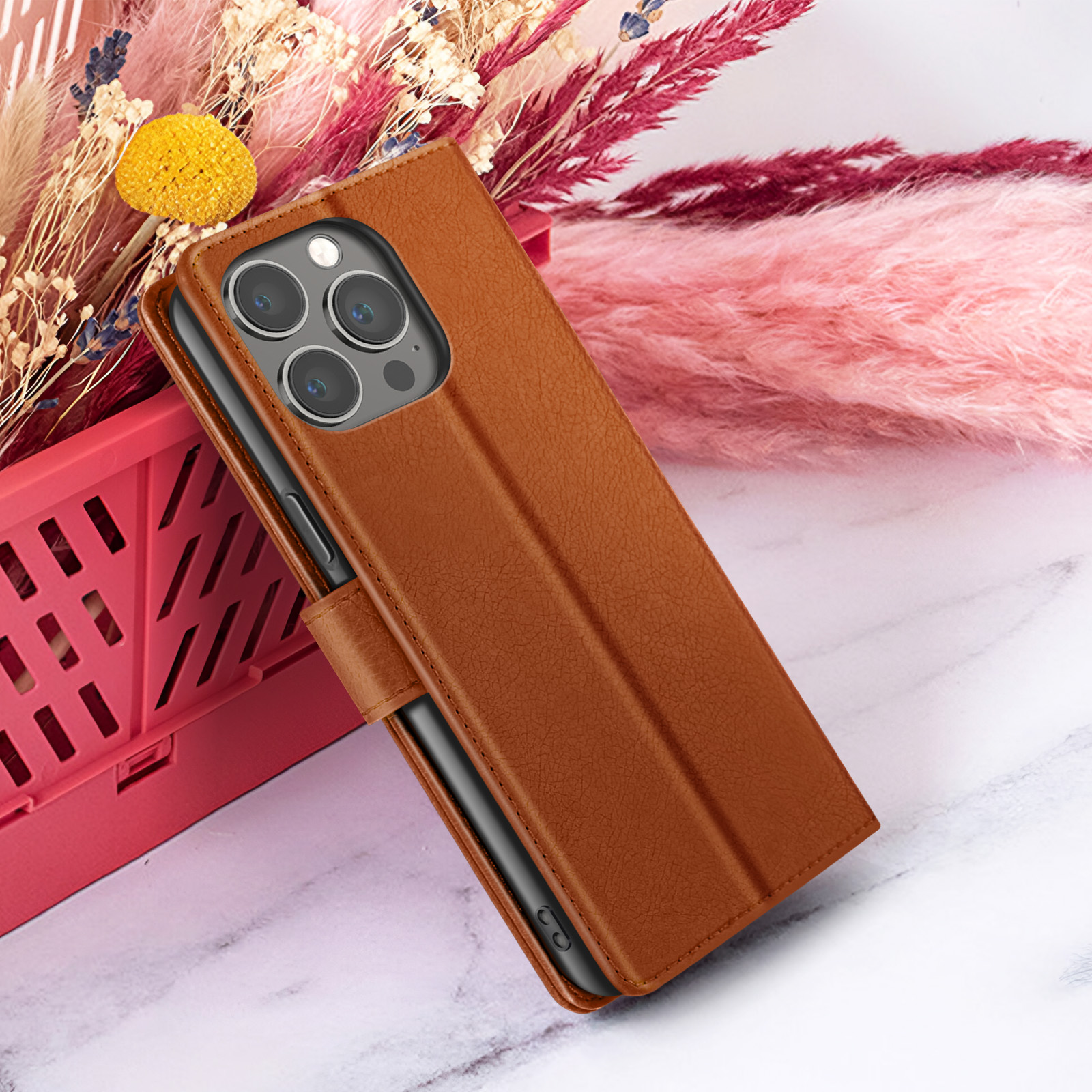 Een bruine leren telefoonhoes met een telefoon erin, tegen een achtergrond van roze bloemen en een marmeren oppervlak.