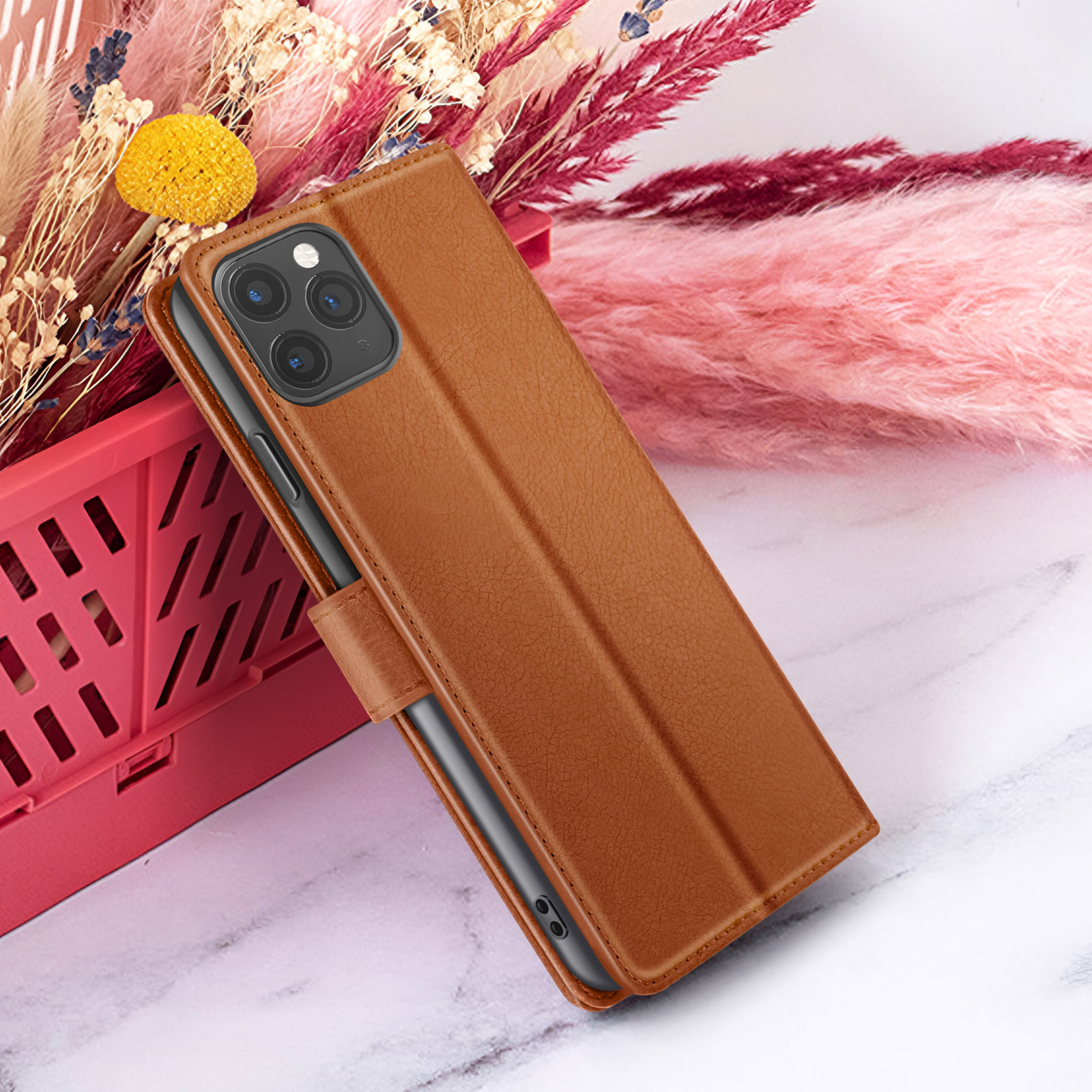 Un étui de téléphone marron avec un téléphone à l'intérieur, adossé à des fleurs séchées. Le fond comprend des éléments roses et blancs.