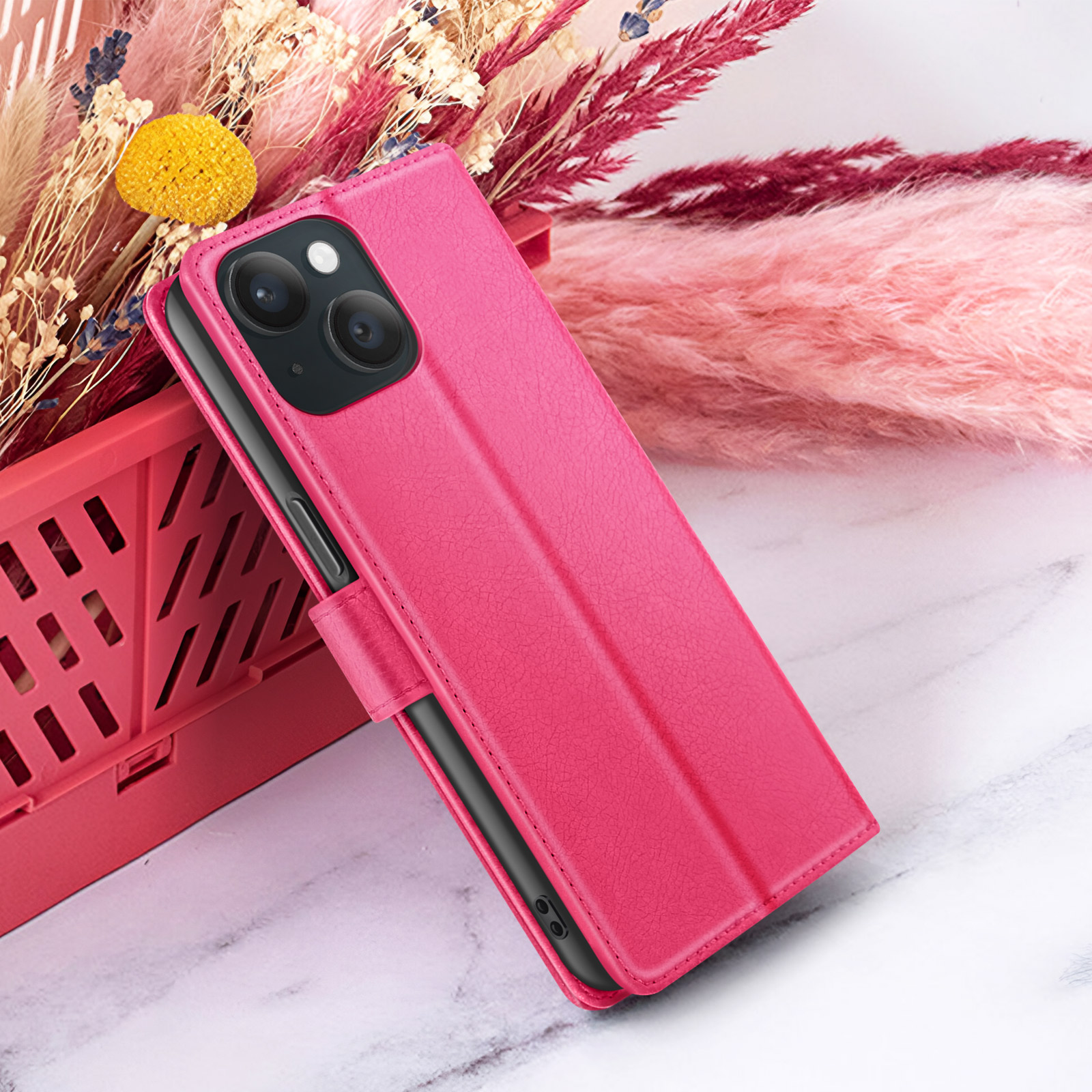 Étui de téléphone rose avec un téléphone à l'intérieur, appuyé contre des fleurs roses et un contenant rouge, sur une surface en marbre.