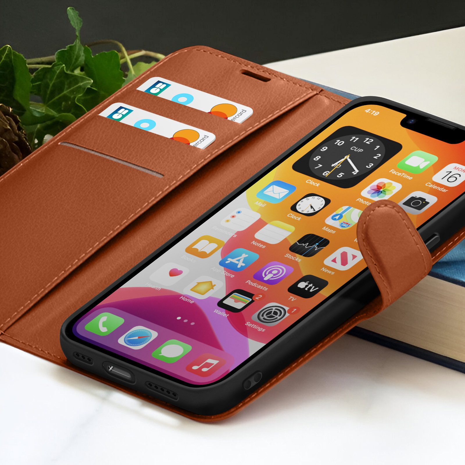 Een telefoon in een bruine hoes geeft apps weer. Twee kaarten zitten in een sleuf. De hoes is open, naast een boek en een groene plant.