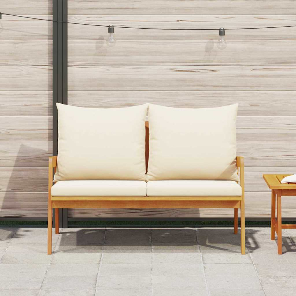 Ein hölzernes Outdoor-Liegesofa mit cremefarbenen Kissen. Rechts steht ein kleiner Tisch. Lichterketten hängen darüber. Holzwand im Hintergrund.