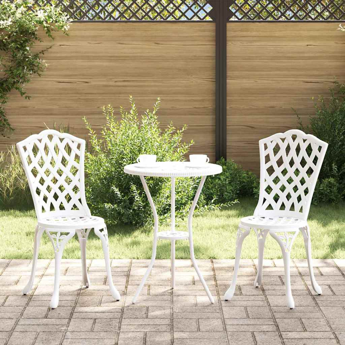 Weißes Bistro-Set im Freien auf einer Terrasse. Beinhaltet zwei Stühle und einen kleinen Tisch mit zwei Tassen. Grüne Büsche und Holzzaun im Hintergrund.
