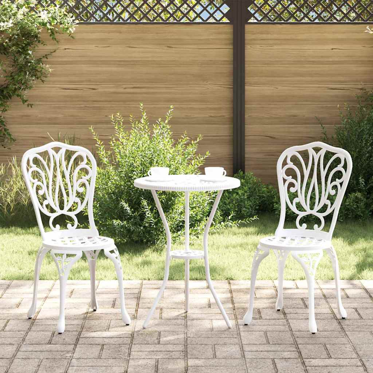 Juego de patio blanco, dos sillas y una mesa pequeña con tazas, en un patio de ladrillo. El fondo muestra una valla de madera y plantas verdes.