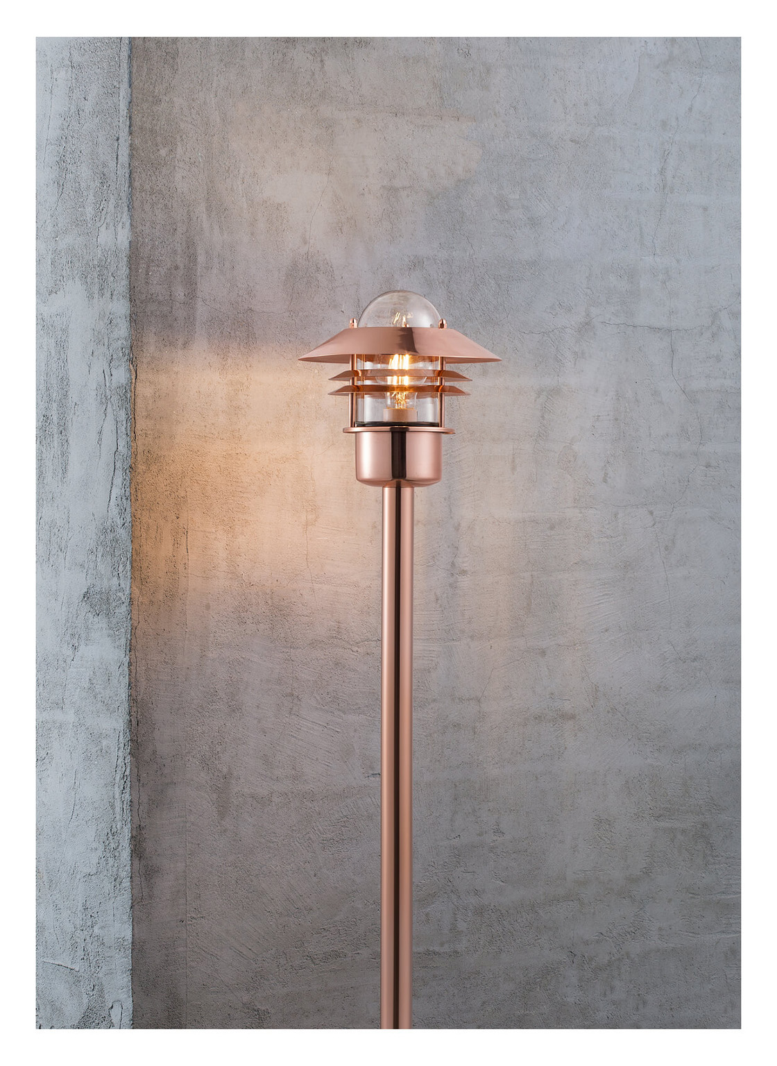 Eine kupferfarbene Außenlampe mit einem klaren Glaskolben und einem mehrschichtigen Schirm steht vor einer Betonmauer. Das Licht ist an.