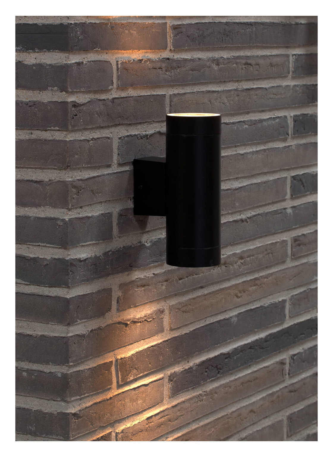 Eine schwarze zylindrische Außenwandleuchte an einer grauen Backsteinmauer. Die Lampe strahlt warmes Licht nach oben und unten aus.