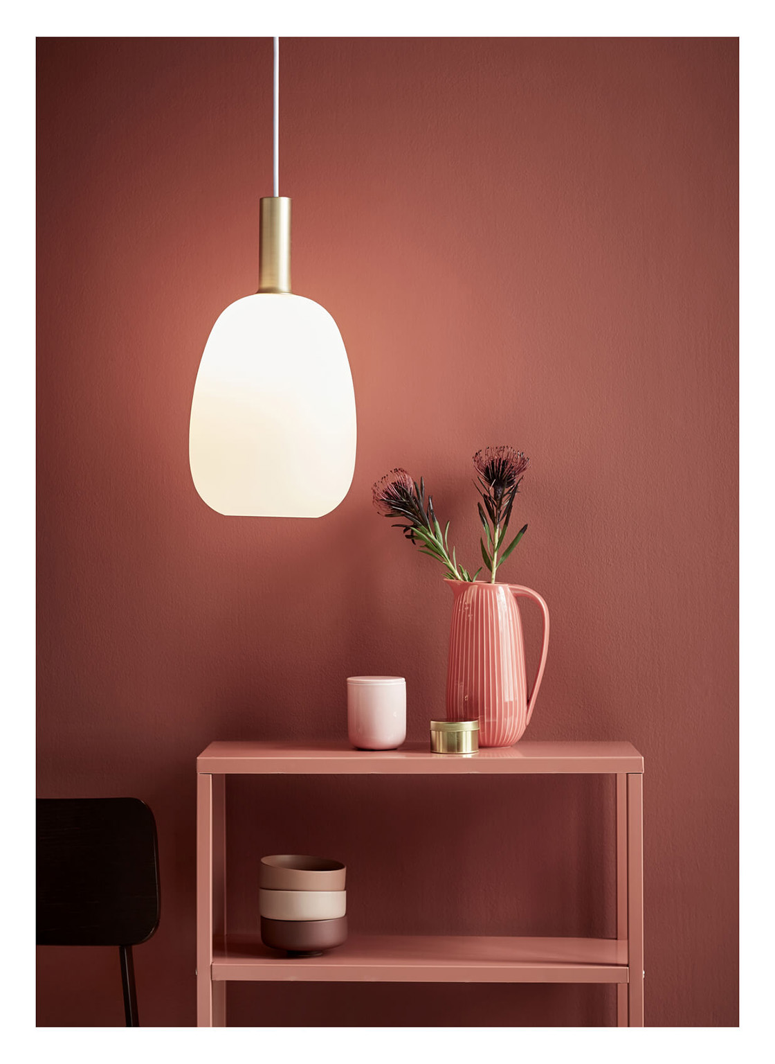 Une suspension lumineuse est suspendue au-dessus d'une étagère rose avec un vase de fleurs, une tasse et des bols décoratifs. Une chaise sombre est visible.