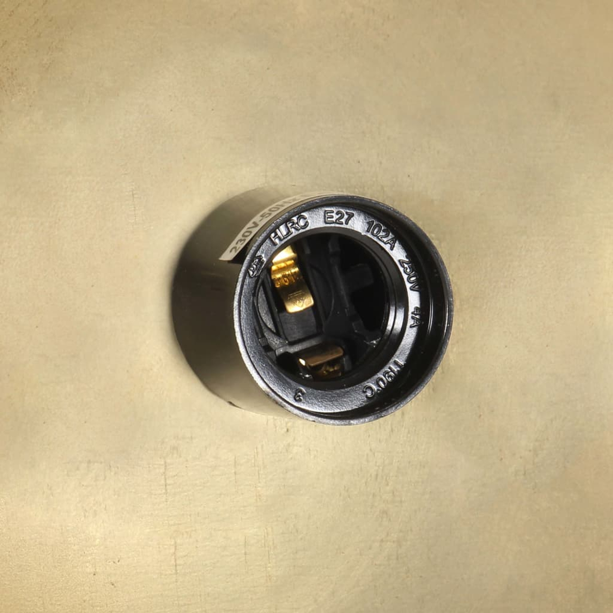 Eine schwarze E27-Glühbirnenfassung mit goldenen Kontaktpunkten auf einer beigefarbenen Oberfläche, verschiedene Markierungen.