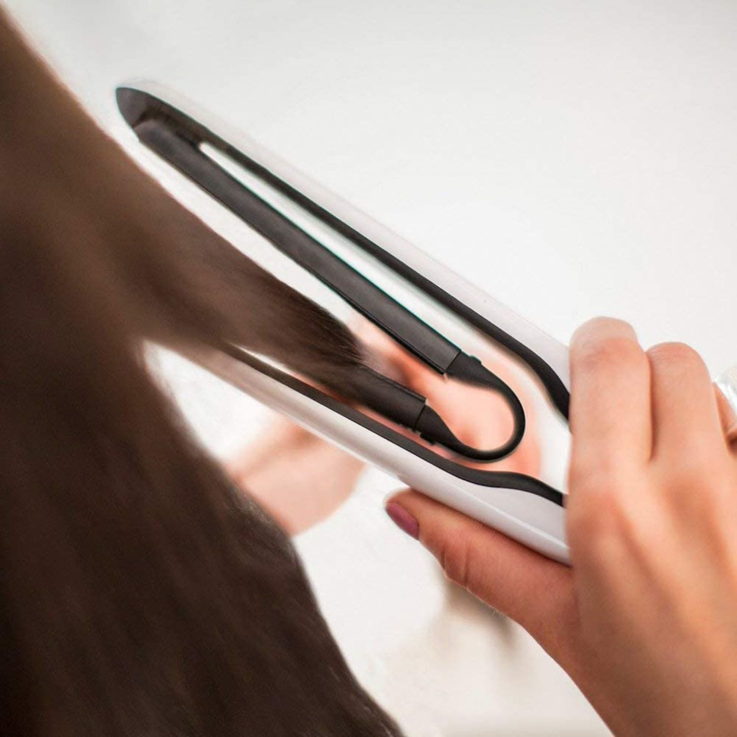 Eine Person, die einen Haarglätter auf braunem Haar verwendet. Der Glätter ist weiß und schwarz. Hand, die den Glätter hält.