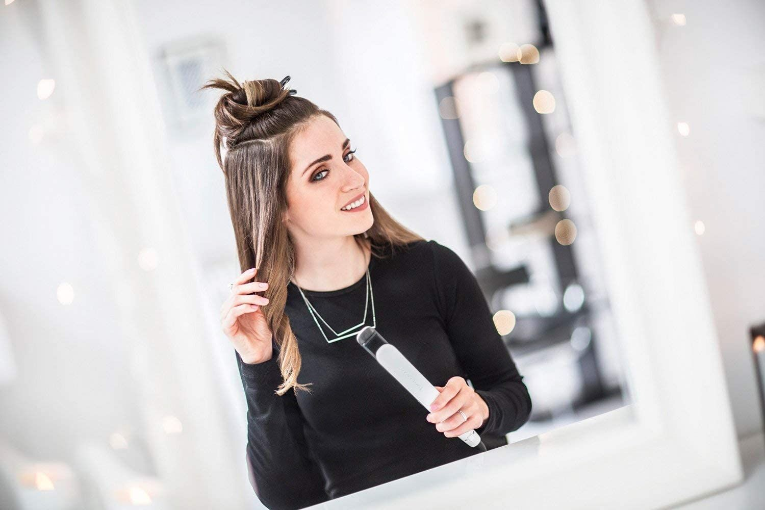 Eine Frau mit braunen Haaren stylt ihr Haar mit einem Glätteisen. Sie trägt ein schwarzes Hemd und eine silberne Halskette.