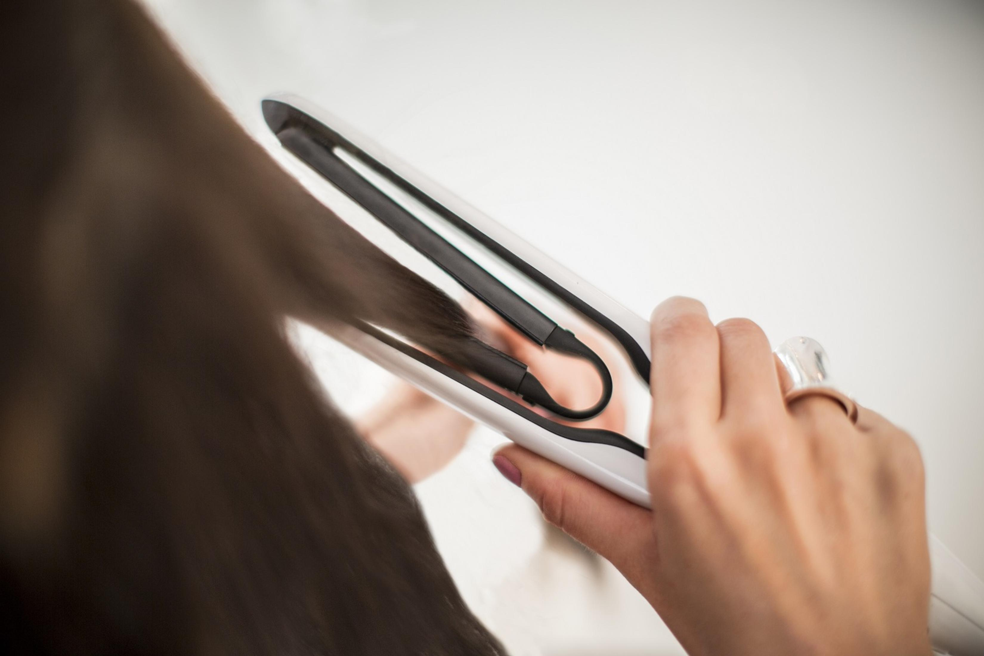 Eine Person, die einen weißen und schwarzen Haarglätter an langen braunen Haaren verwendet, mit einer be-ringten Hand im Vordergrund.