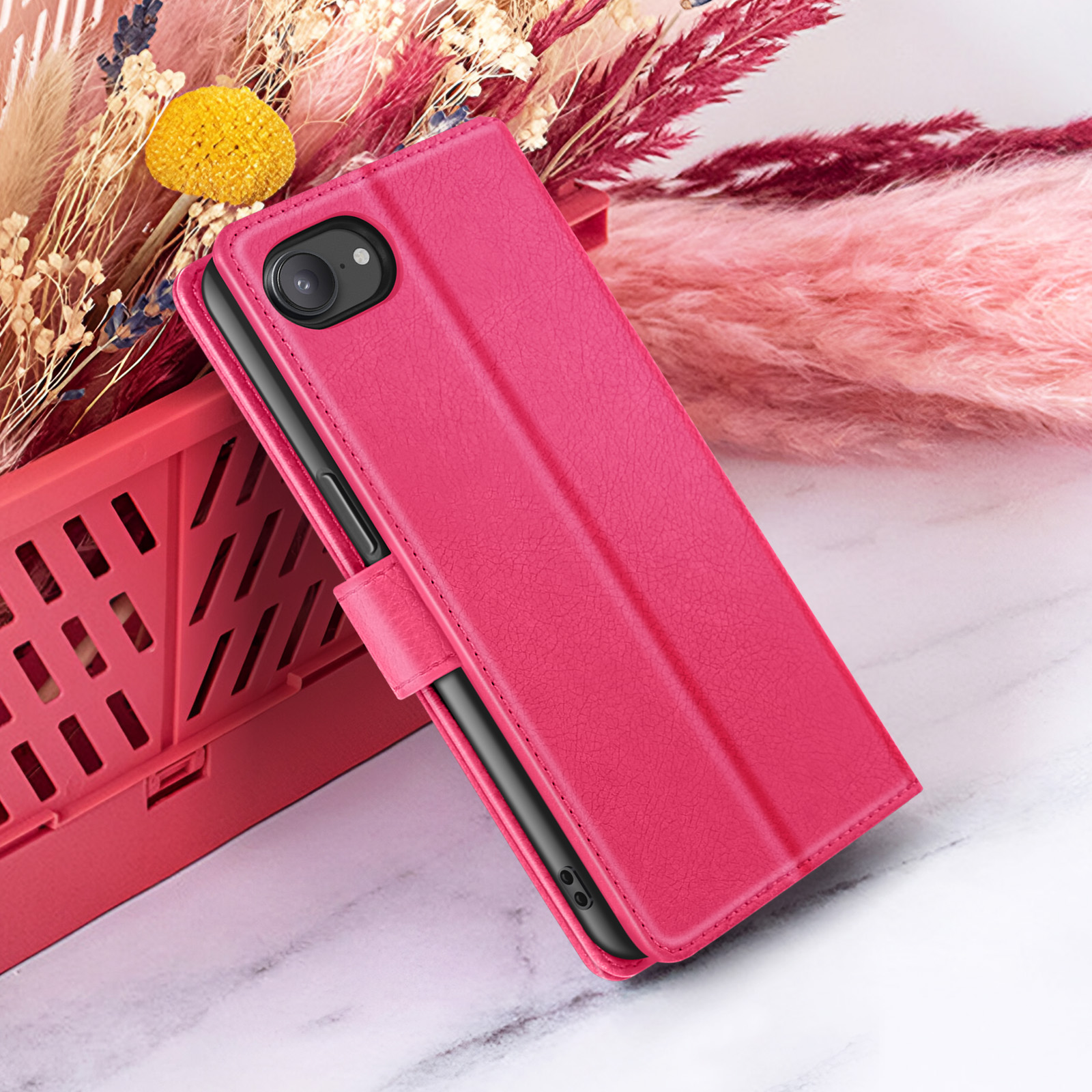 Een roze telefoonhoesje met een telefoon erin rust op een roze mand met bloemen op de achtergrond, op een marmeren oppervlak.