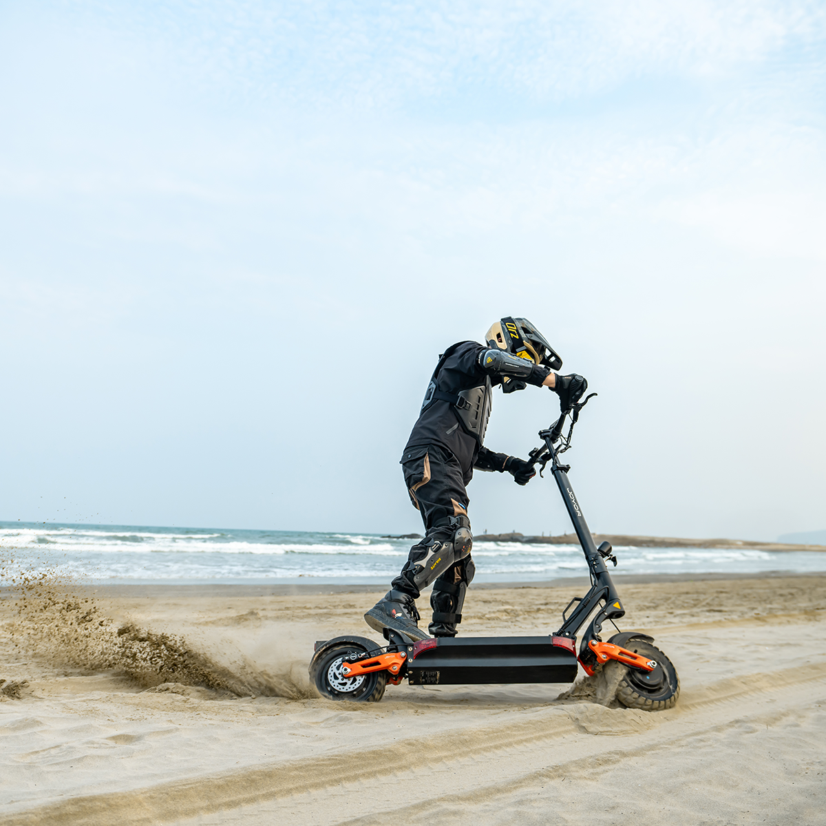Eine Person in Schutzkleidung fährt mit einem Elektroroller an einem Sandstrand. Das Meer ist im Hintergrund unter einem bewölkten Himmel.