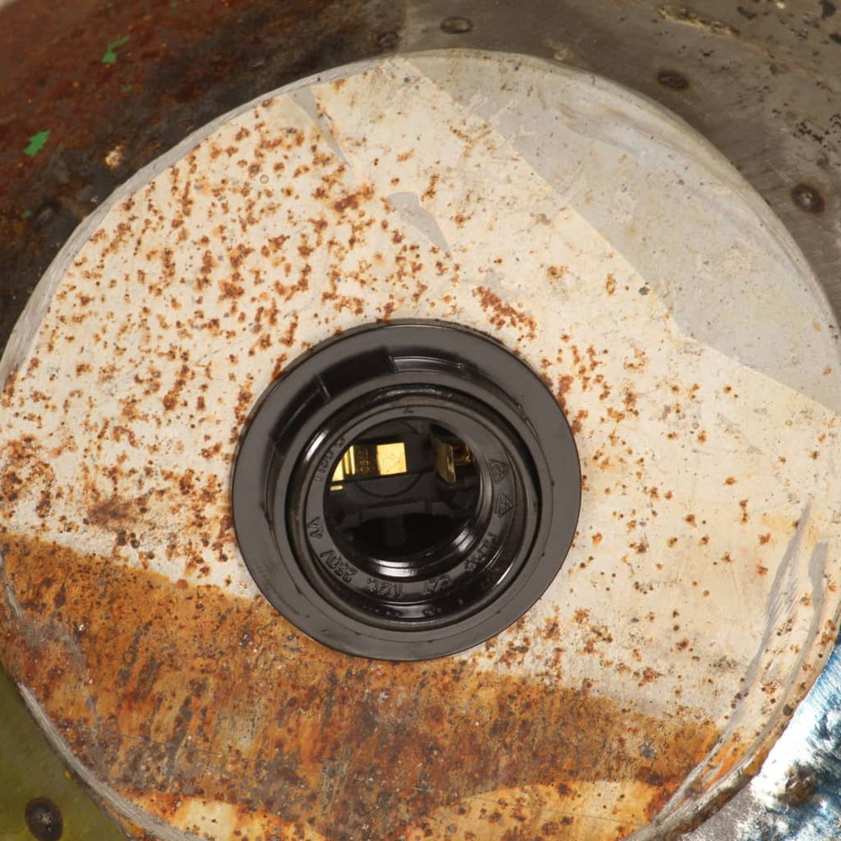 Nahaufnahme eines rostigen Metallobjekts mit einer schwarzen, kreisförmigen Fassung in der Mitte. Die Fassung zeigt elektrische Bauteile.