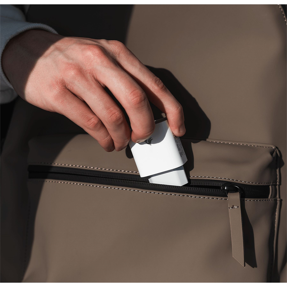 Eine Hand legt ein weißes Ladegerät in eine beige Rucksacktasche. Der Reißverschluss ist schwarz. Der Hintergrund ist unscharf.