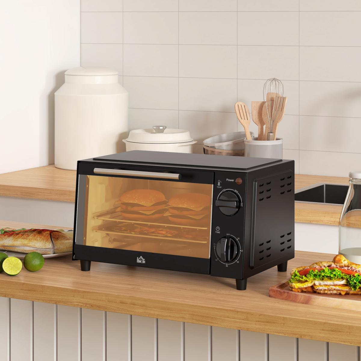 Schwarzer Toasterofen mit zwei Burgern. Limetten und ein Sandwich liegen auf der Theke. Holzbesteck und Gläser sind im Hintergrund.