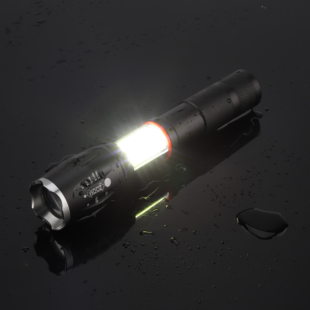 Schwarze Taschenlampe auf nassem, dunklem Untergrund. Das Licht ist an. Die Taschenlampe hat einen Zoom und ein zusätzliches Licht.