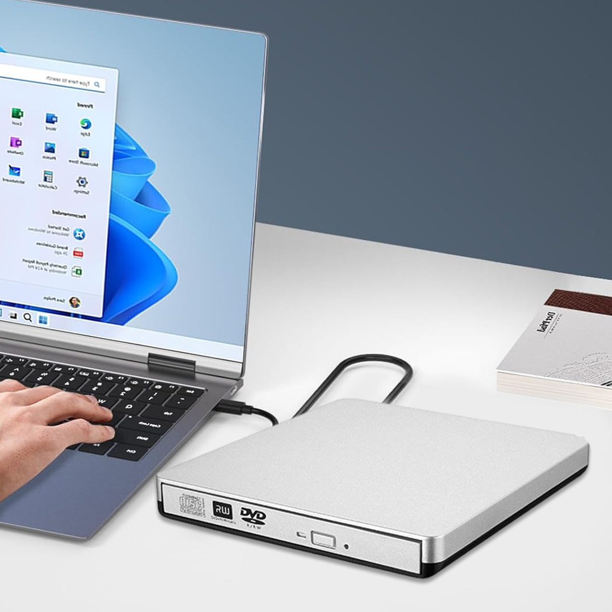 Ein Laptop mit einer Hand auf der Tastatur, verbunden mit einem silbernen DVD-Laufwerk. Ein Buch liegt auf dem Tisch.
