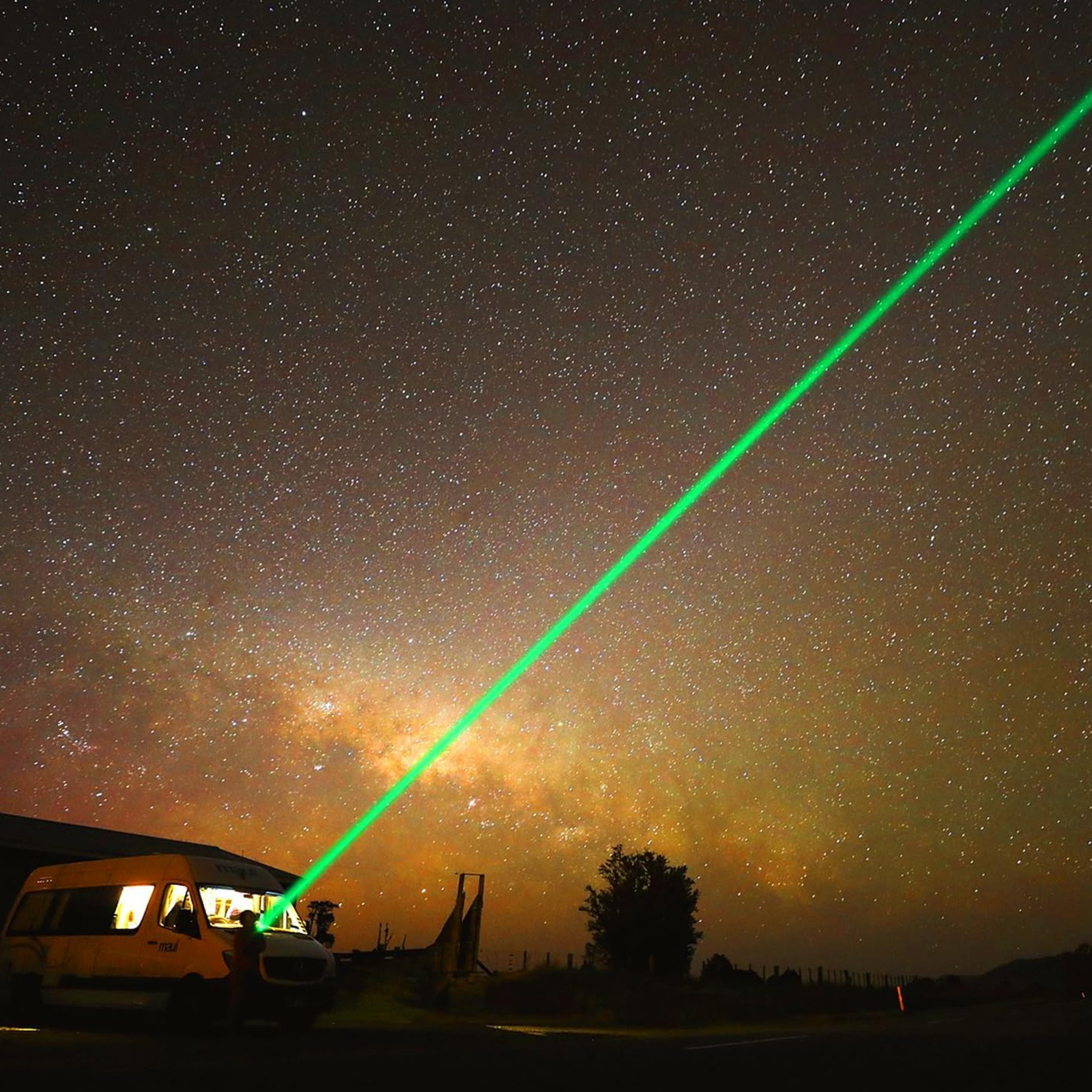 Ein grüner Laserstrahl schießt von einem Van in den sternenklaren Nachthimmel. Die Milchstraße ist sichtbar. Eine Person steht am Van.
