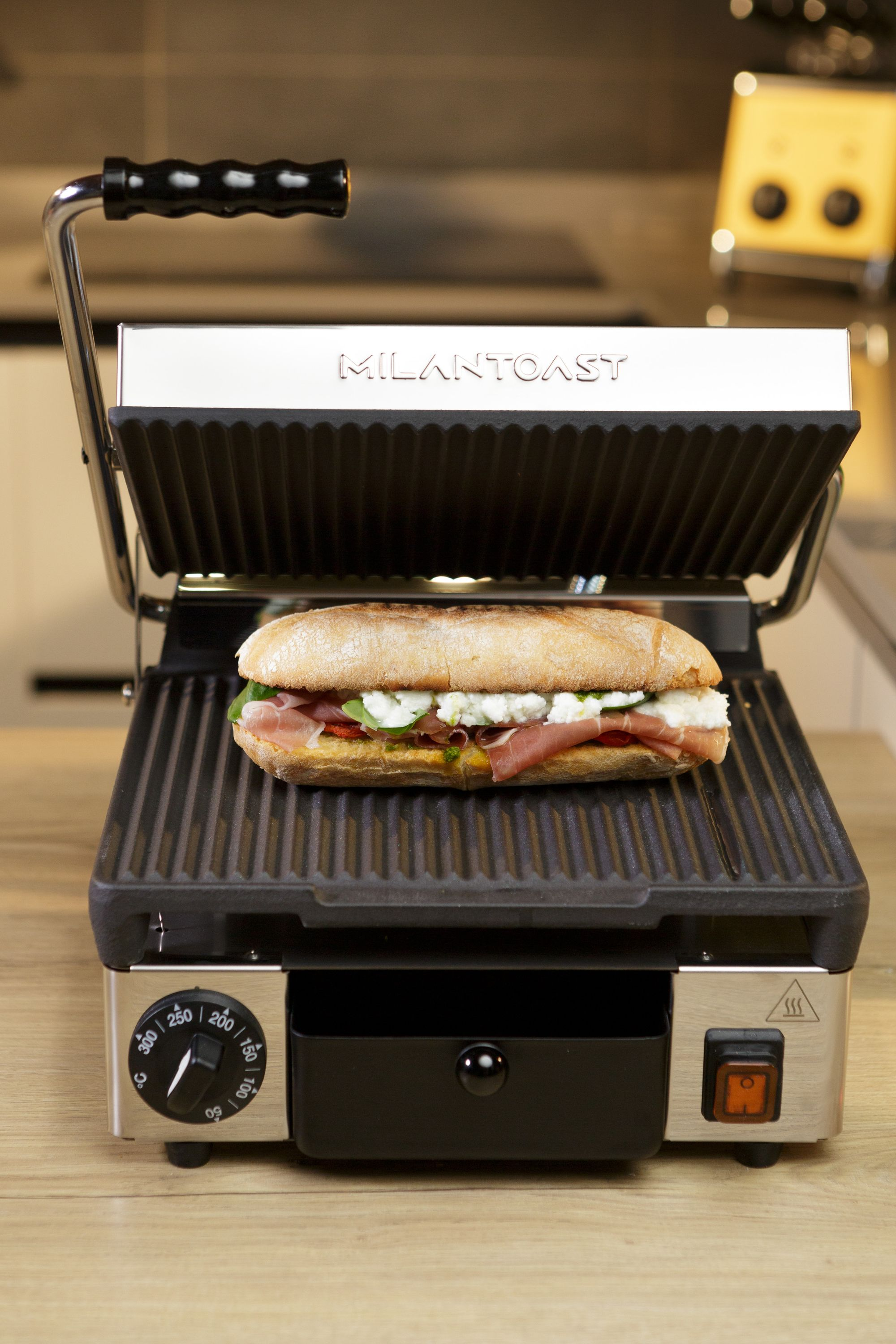 Ein Panini-Grill mit einem Sandwich darin. Das Sandwich hat Fleisch, Käse und Pesto. Die Presse steht auf einem Holztisch.