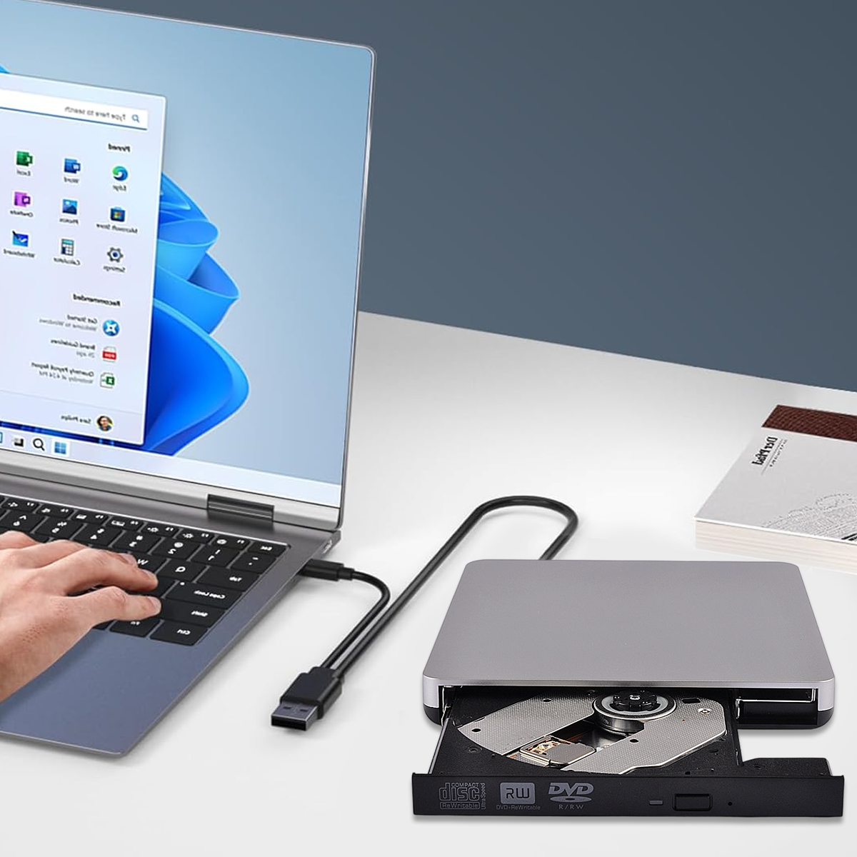 Laptop mit silbernem DVD-Laufwerk verbunden. Eine Hand bedient den Laptop. Ein Buch liegt auf dem Tisch.