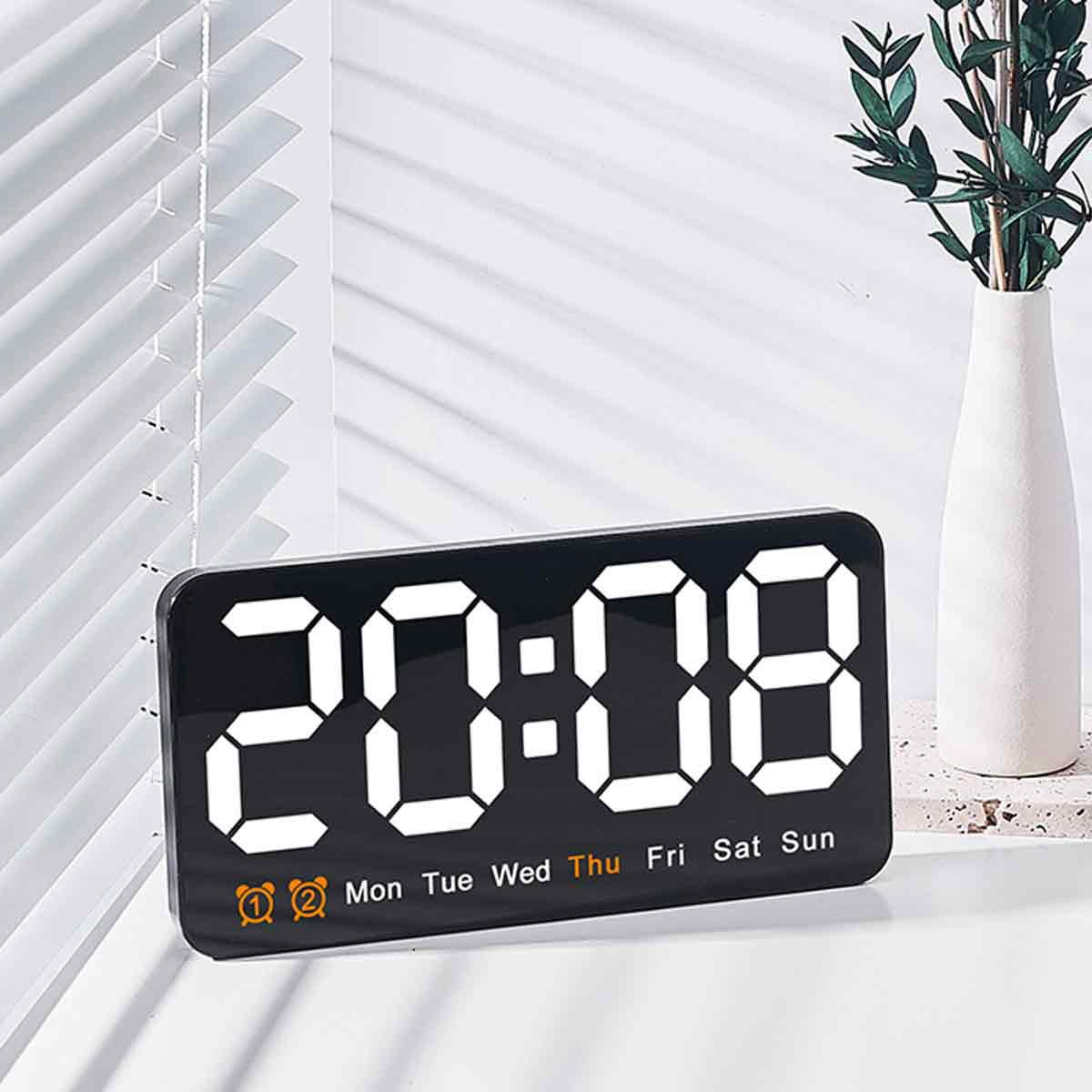 Digitaluhr mit der Anzeige 20:08. Weiße Zahlen auf schwarzem Hintergrund. Eine Vase mit grünen Blättern befindet sich auf der rechten Seite.