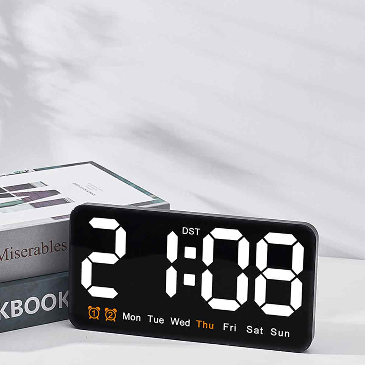Eine schwarze Digitaluhr zeigt 2:08 Uhr an. Im Hintergrund sind Bücher und eine weiße Oberfläche zu sehen. Die Uhr zeigt auch den Wochentag an.