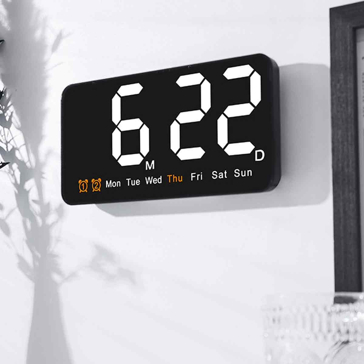 Schwarze rechteckige Digitaluhr zeigt 6:22 an. Sie ist an einer weißen Wand montiert. Wochentage stehen unter der Uhrzeit.