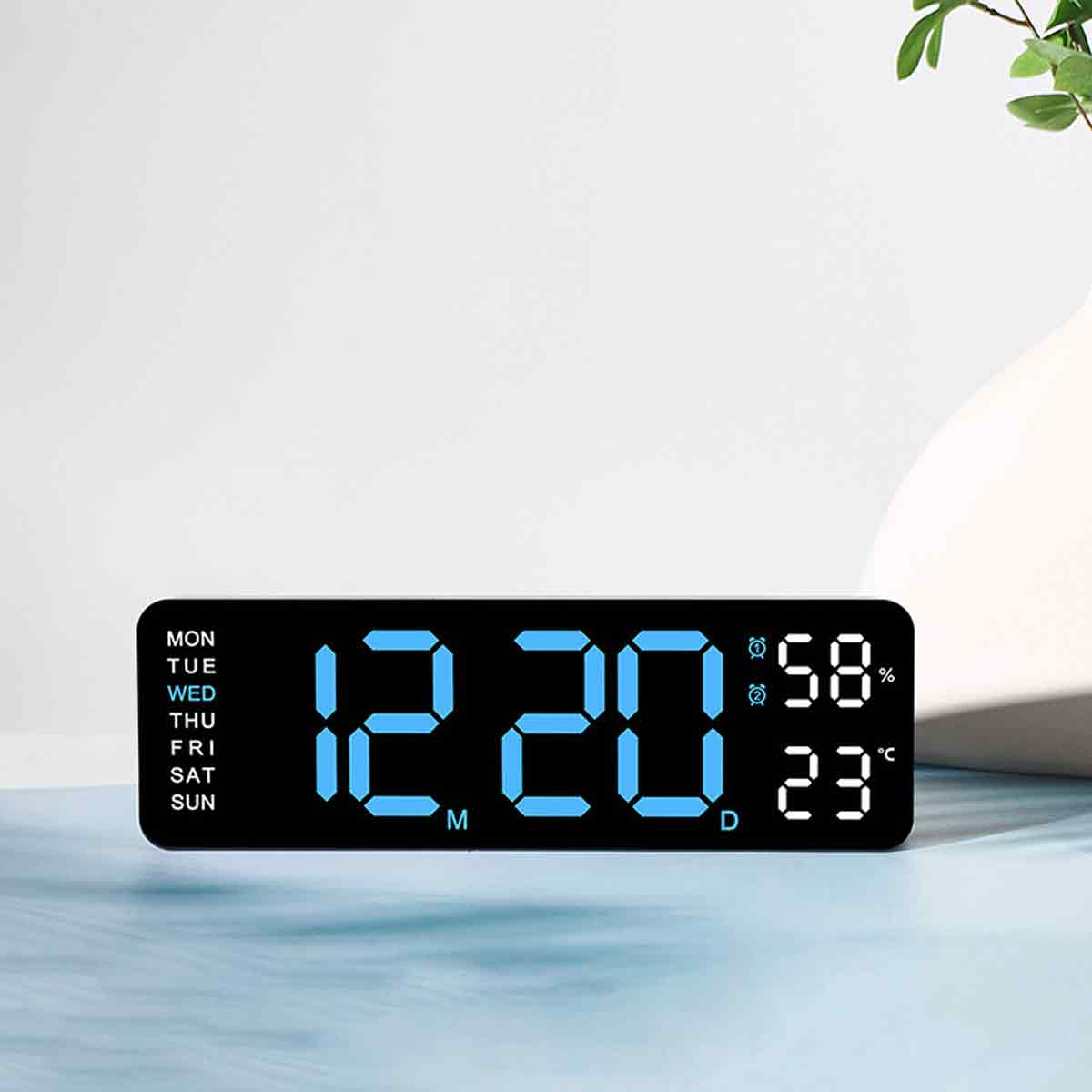 Eine Digitaluhr zeigt 12:20 Uhr mit Feuchtigkeits- und Temperaturanzeigen an. Die Uhr steht auf einer Oberfläche, im Hintergrund eine weiße Vase und grüne Blätter.