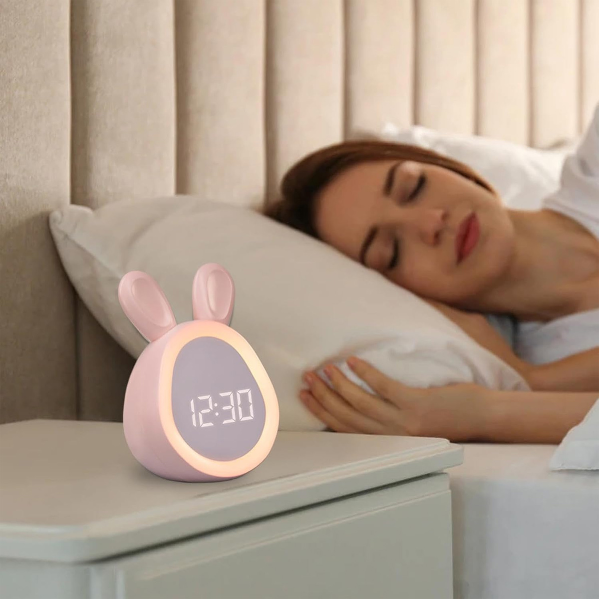 Ein rosa, hasenförmiger Wecker mit der Anzeige 12:30 steht auf einem Nachttisch. Dahinter schläft eine Frau im Bett.