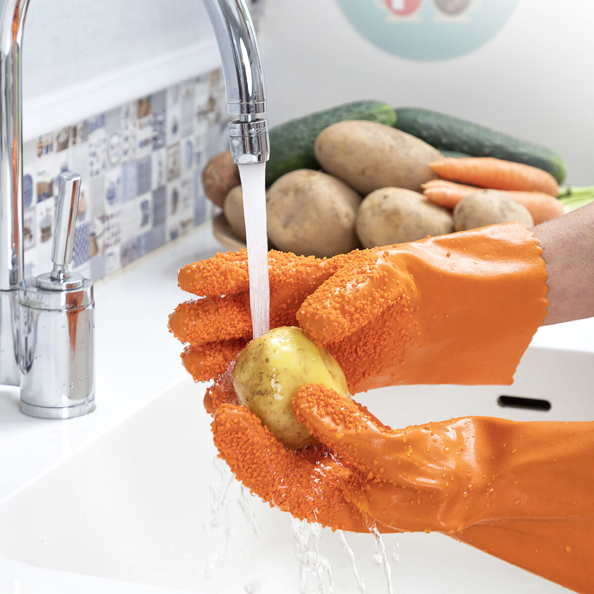 Guanti arancioni lavano una patata sotto l'acqua corrente. Sullo sfondo verdure come patate, cetrioli e carote.