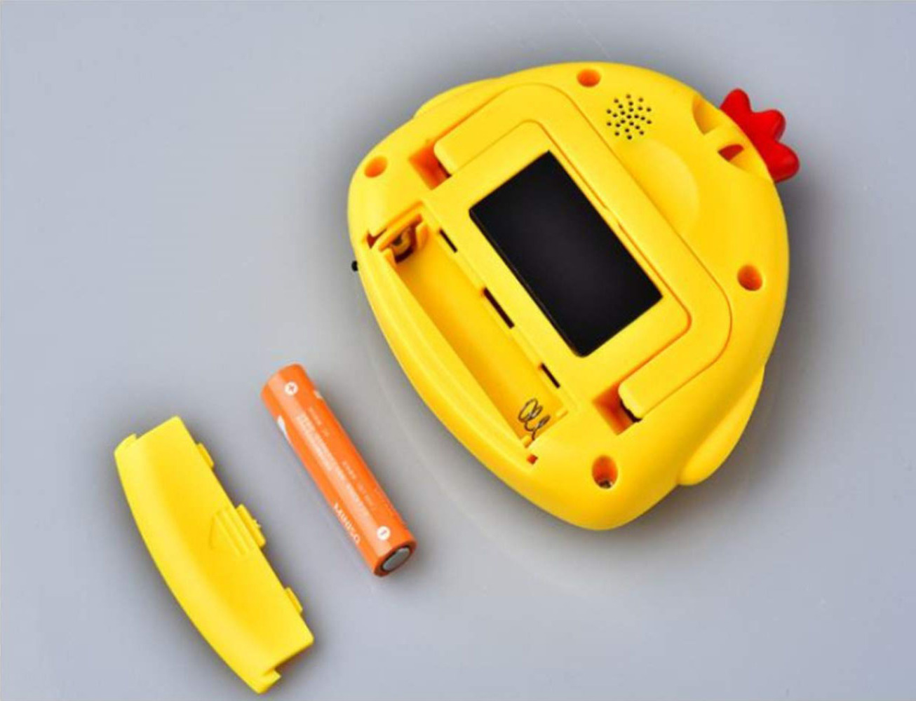 Ein gelbes Spielzeug in Form eines Huhns mit schwarzem Bildschirm, Batteriefach, orangefarbener Batterie und abgenommenem gelben Deckel auf grauem Untergrund.