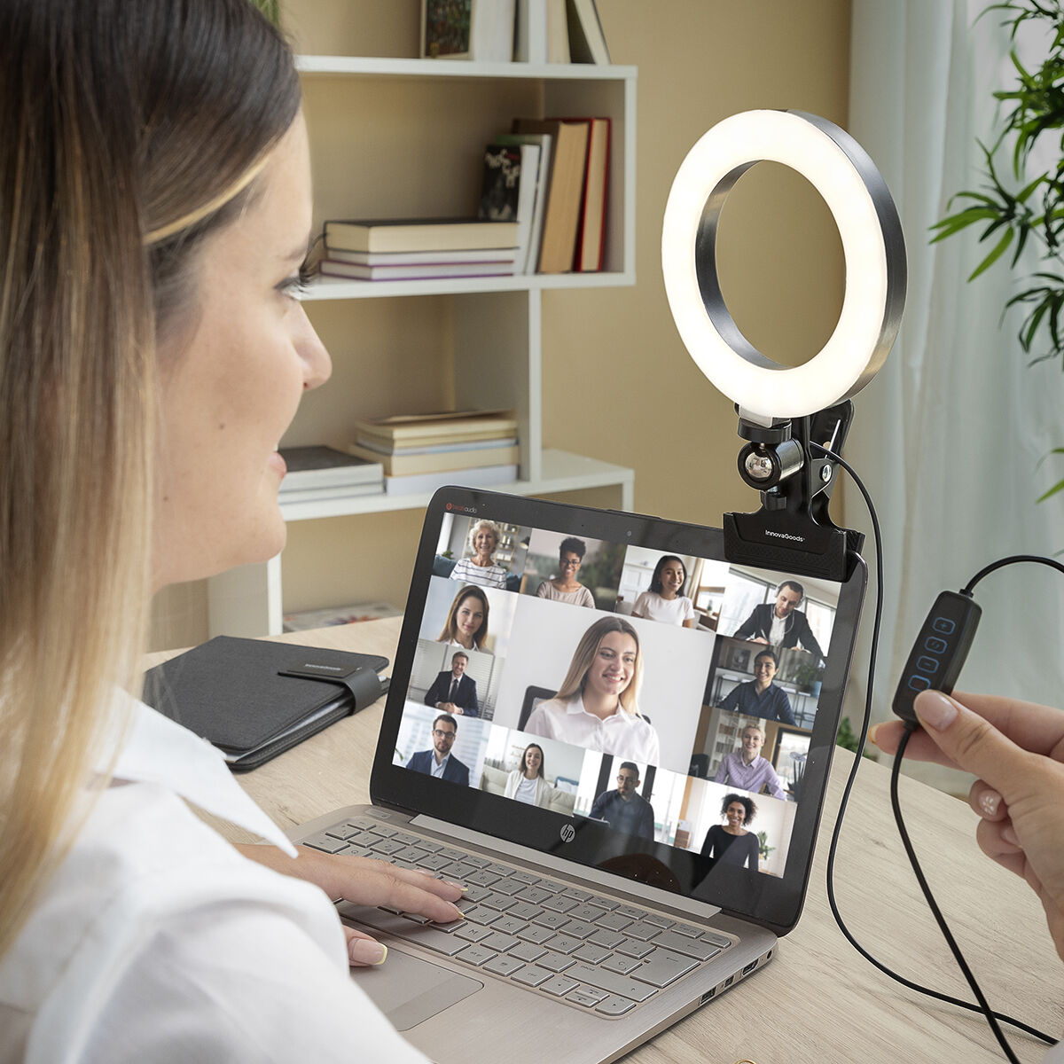 Eine Frau nimmt an einer Videokonferenz auf ihrem Laptop teil. Ein Ringlicht ist angebracht. Eine Hand hält ein Bedienfeld. Bücherregale sind sichtbar.