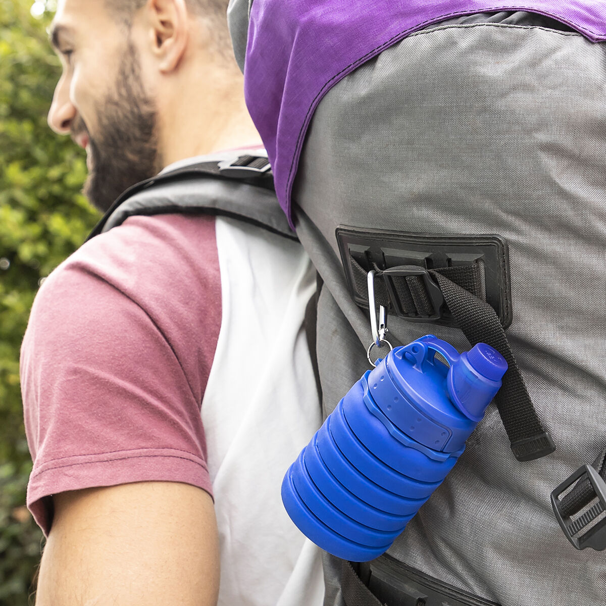 Un uomo con la barba e uno zaino trasporta una borraccia blu pieghevole. Lo zaino è grigio e viola, con cinghie nere.