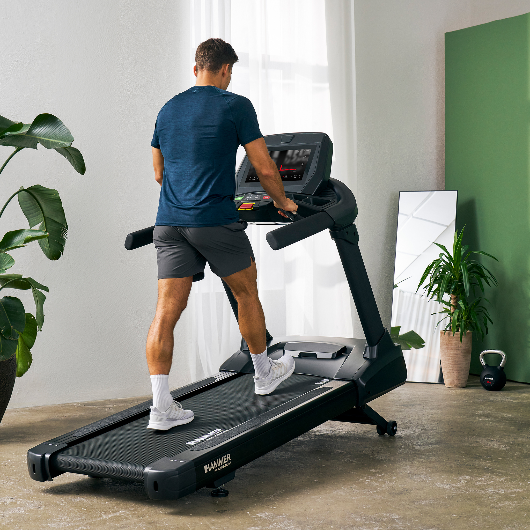 Mann auf Laufband. Zimmer mit Pflanzen, Spiegel und Kettlebell. Er trägt blaues Shirt, graue Shorts, weiße Schuhe. Schwarzes Laufband.
