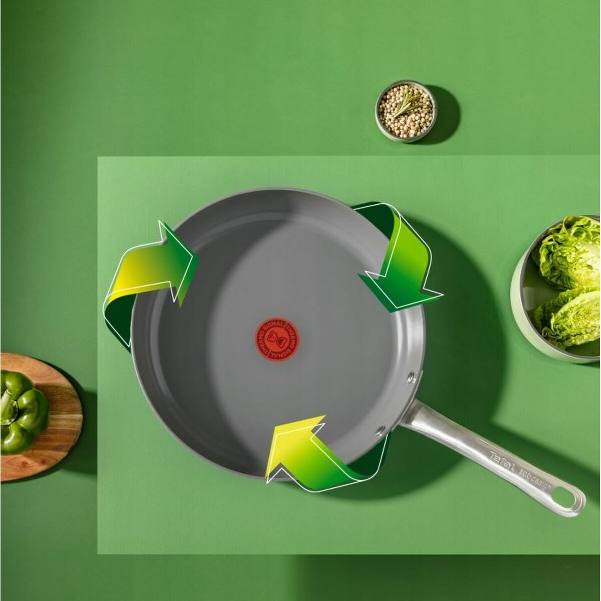 Eine graue Bratpfanne mit einem Recycling-Symbol, umgeben von grünem Gemüse, einer grünen Paprika und einer Schüssel mit Gewürzen auf grünem Hintergrund.