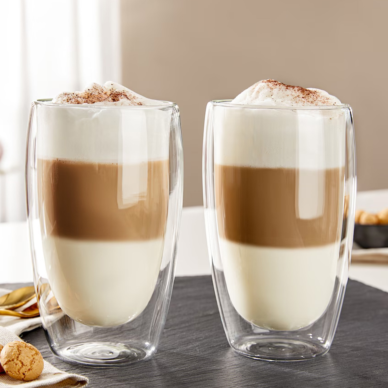 Zwei geschichtete Lattes in klaren Gläsern. Die Schichten sind weiß, braun und mit Schaum und Kakao bedeckt. Ein Keks und ein goldener Löffel sind dabei.