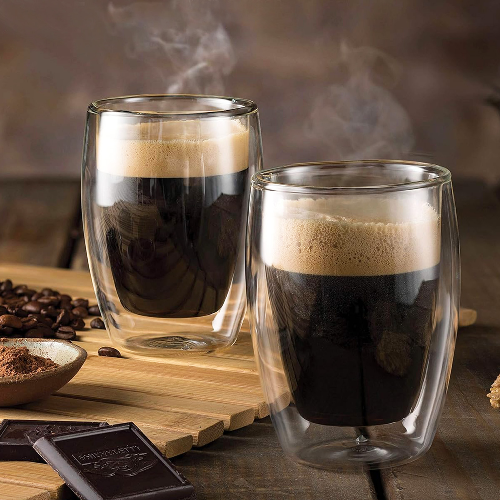 Zwei Glasbecher Kaffee mit Schaum. Schokoladenriegel und Kaffeebohnen auf Holz. Dampf steigt auf.