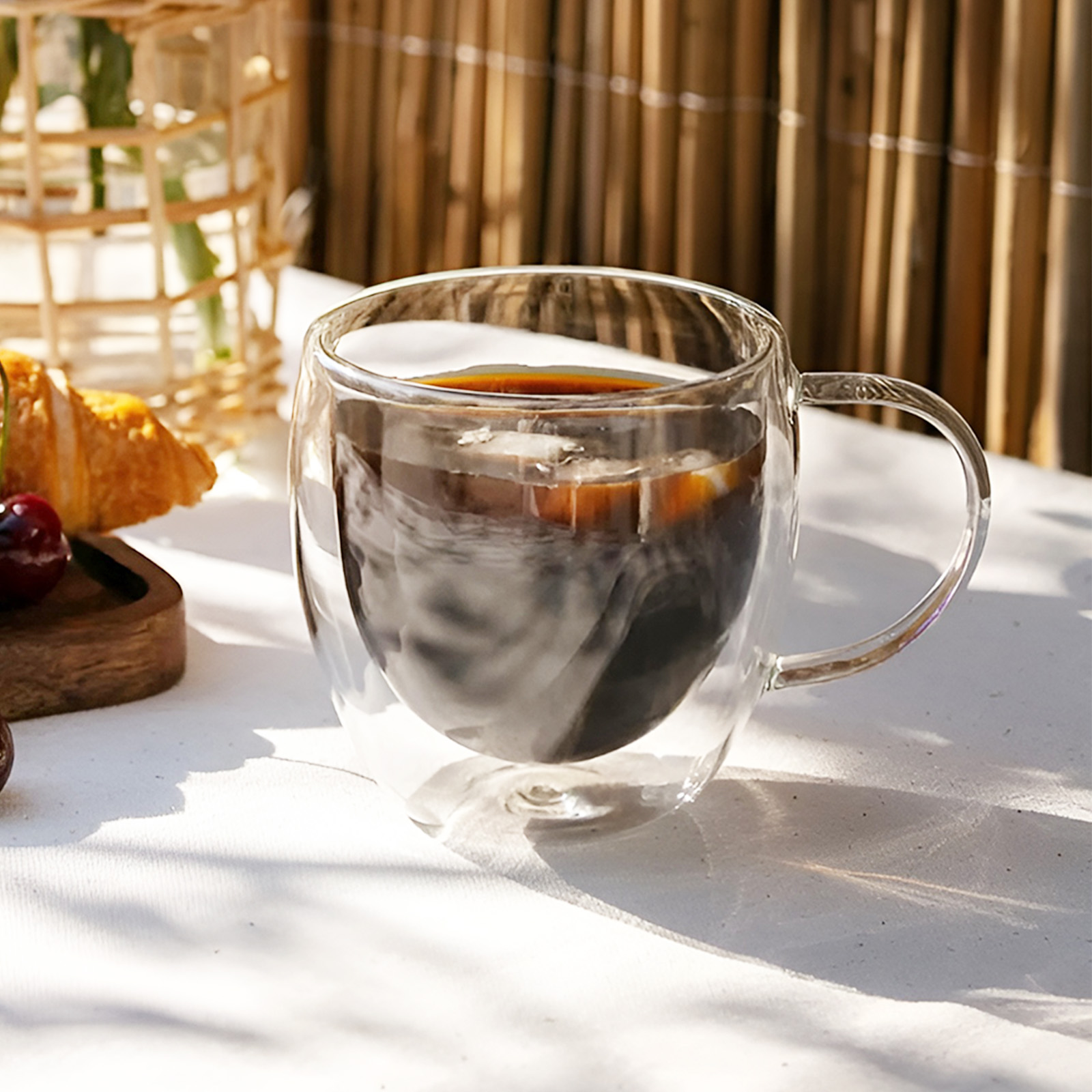 Ein Glasbecher mit dunklem Kaffee und Eiswürfeln auf einer weißen Oberfläche, mit Croissant und Kirschen.