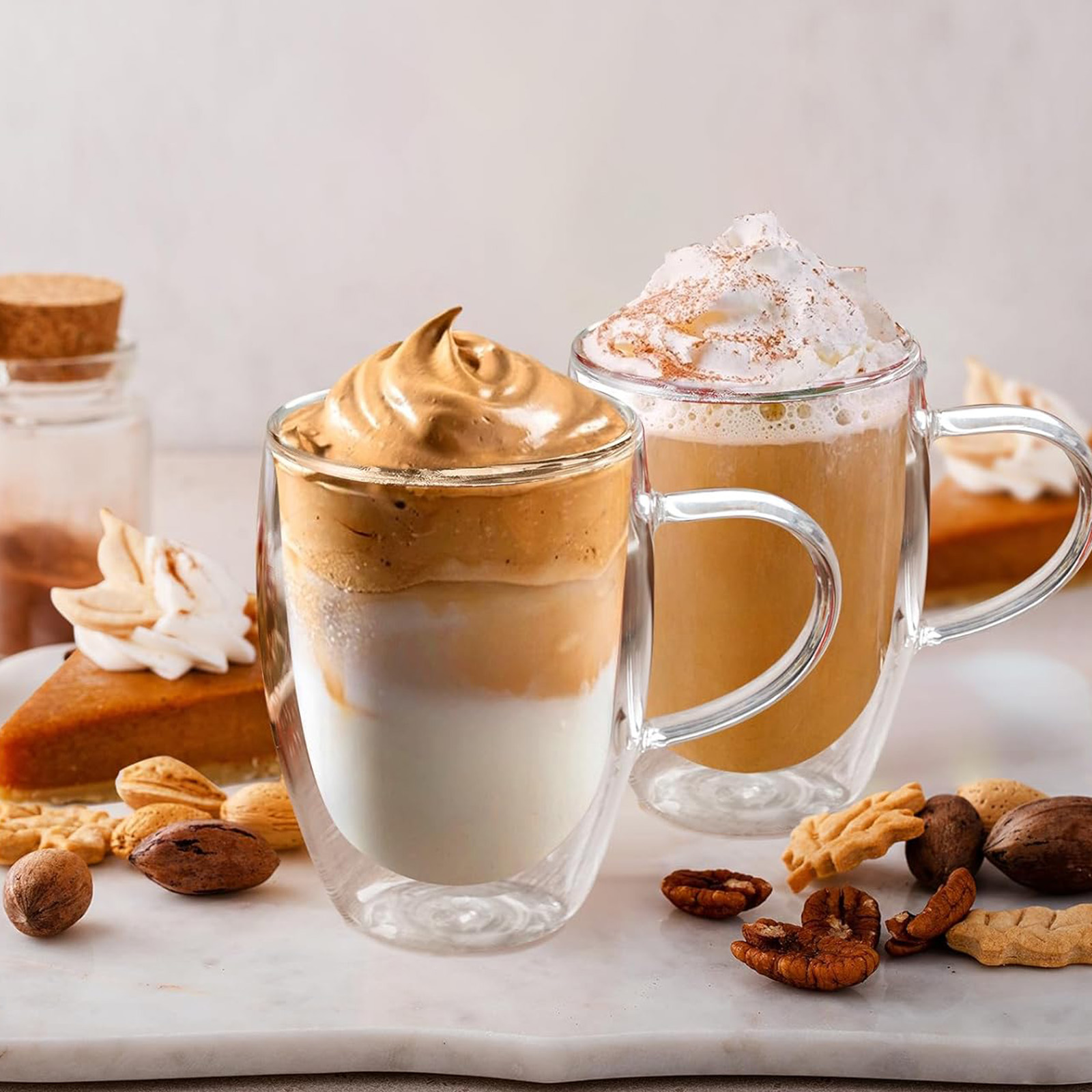 Zwei Kaffeegetränke, eines mit geschlagenem Schaum und das andere mit Schlagsahne. Kürbiskuchenstücke, Nüsse und Kekse liegen auf einer Marmoroberfläche.