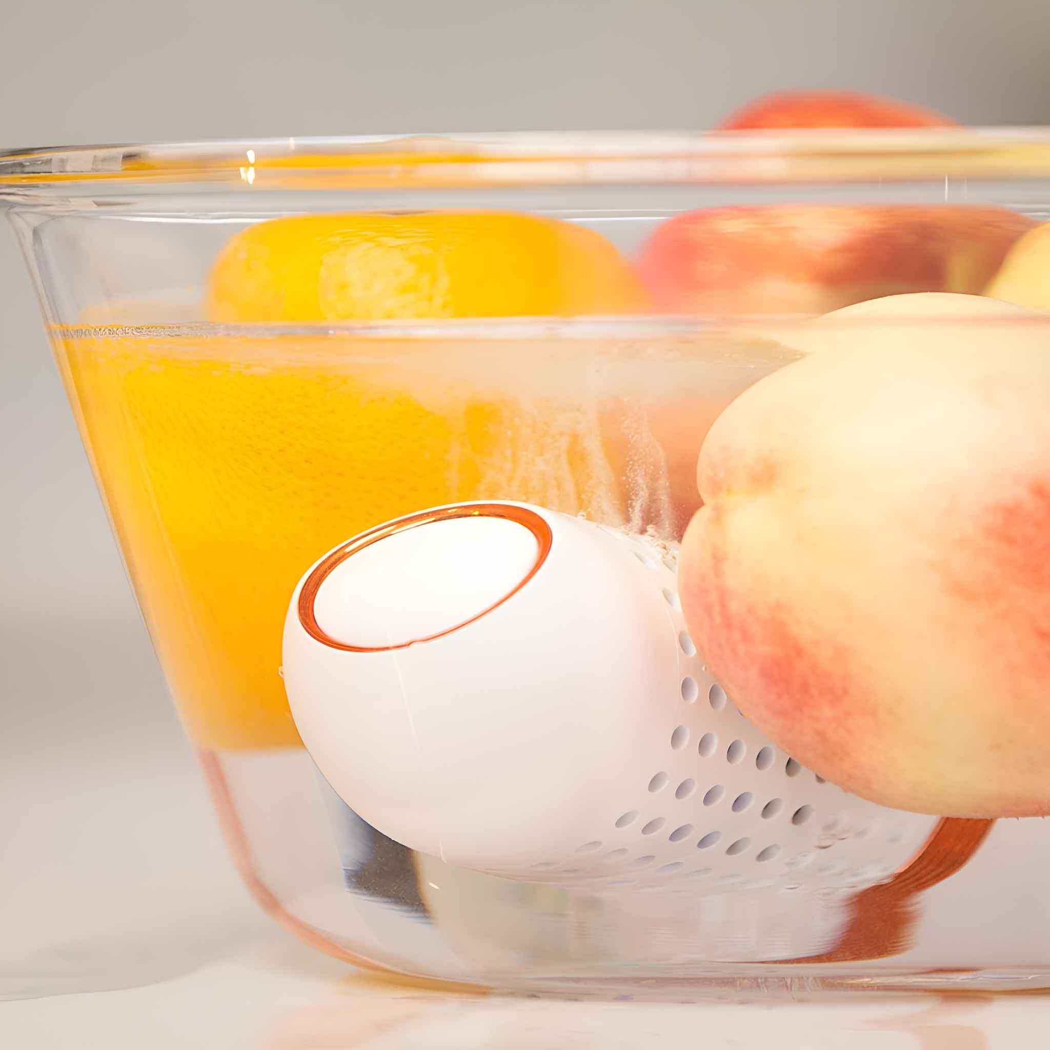 Eine klare Glasschüssel mit Wasser, Früchten und einem weißen Gerät. Orangen und Pfirsiche sind sichtbar. Das Gerät ist weiß mit einem orangenen Ring.
