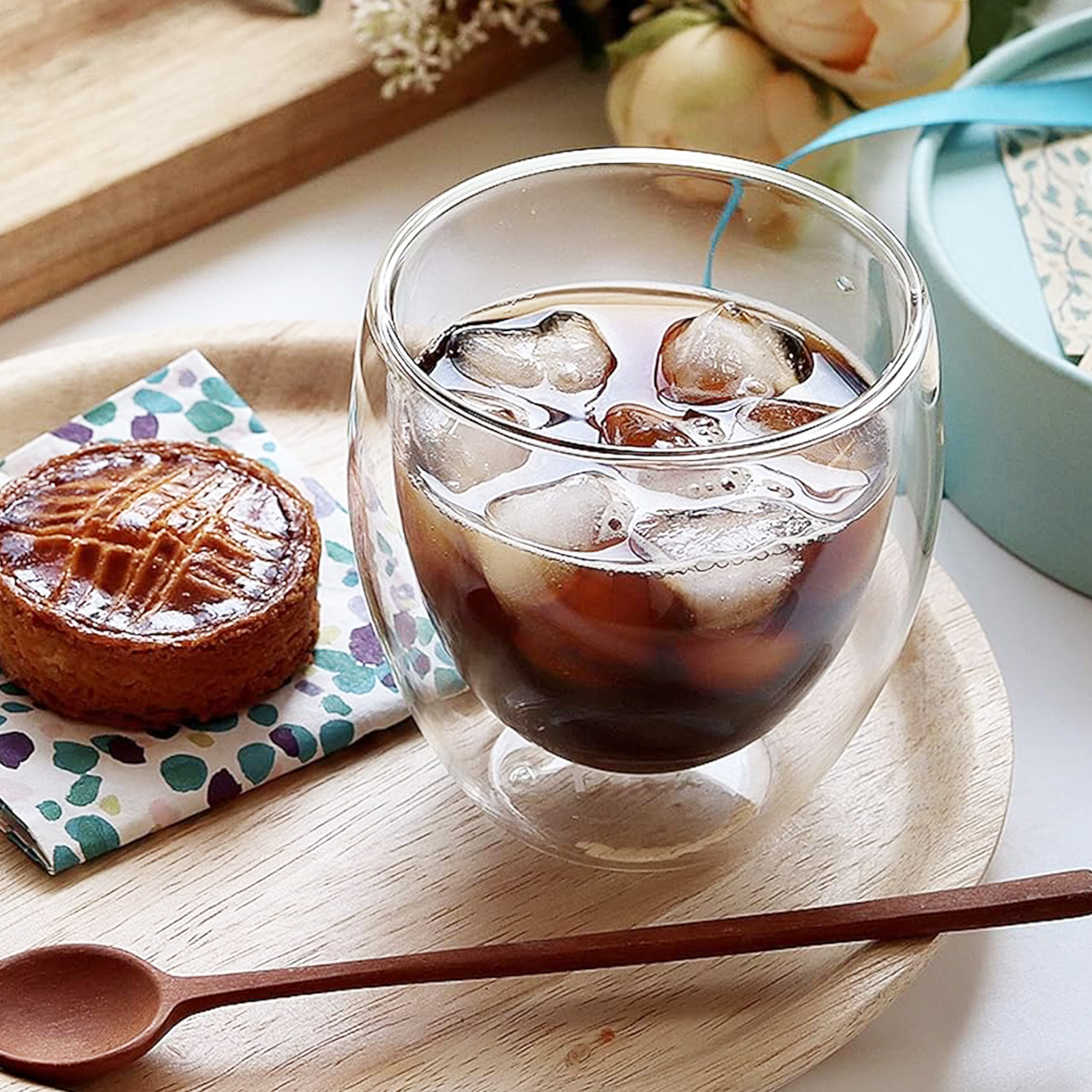 Ein Glas Eiskaffee mit Kuchen, ein Holzlöffel und eine Geschenkbox auf einem Holztablett. Blumen im Hintergrund.