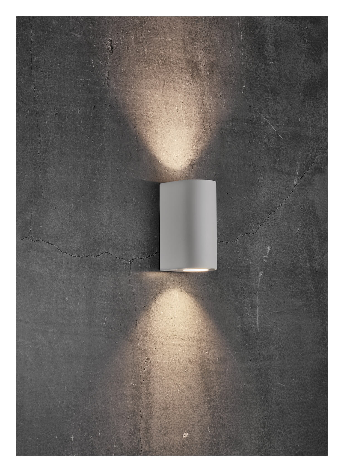 Eine weiße, zylindrische Wandleuchte ist an einer strukturierten, grauen Wand befestigt. Die Lampe wirft ein warmes Licht nach oben und unten.