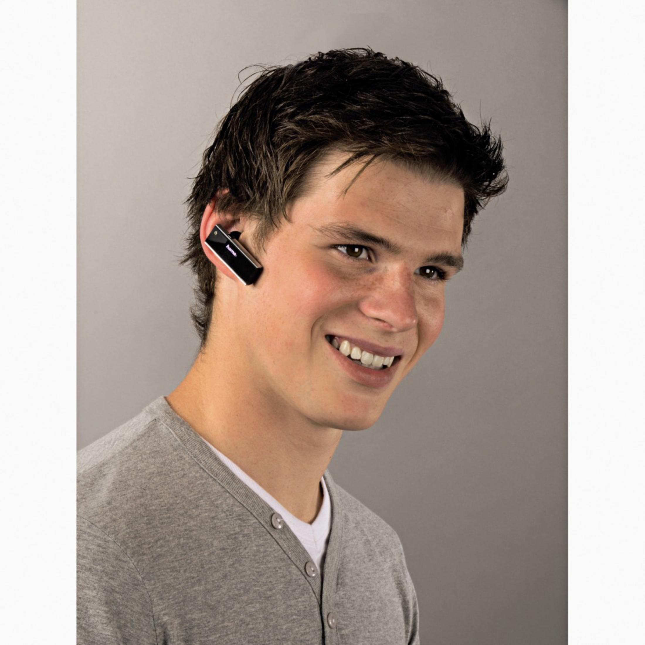 Lächelnder junger Mann mit Bluetooth-Headset. Er hat dunkles Haar, helle Haut und einen grauen Pullover über einem weißen Hemd.