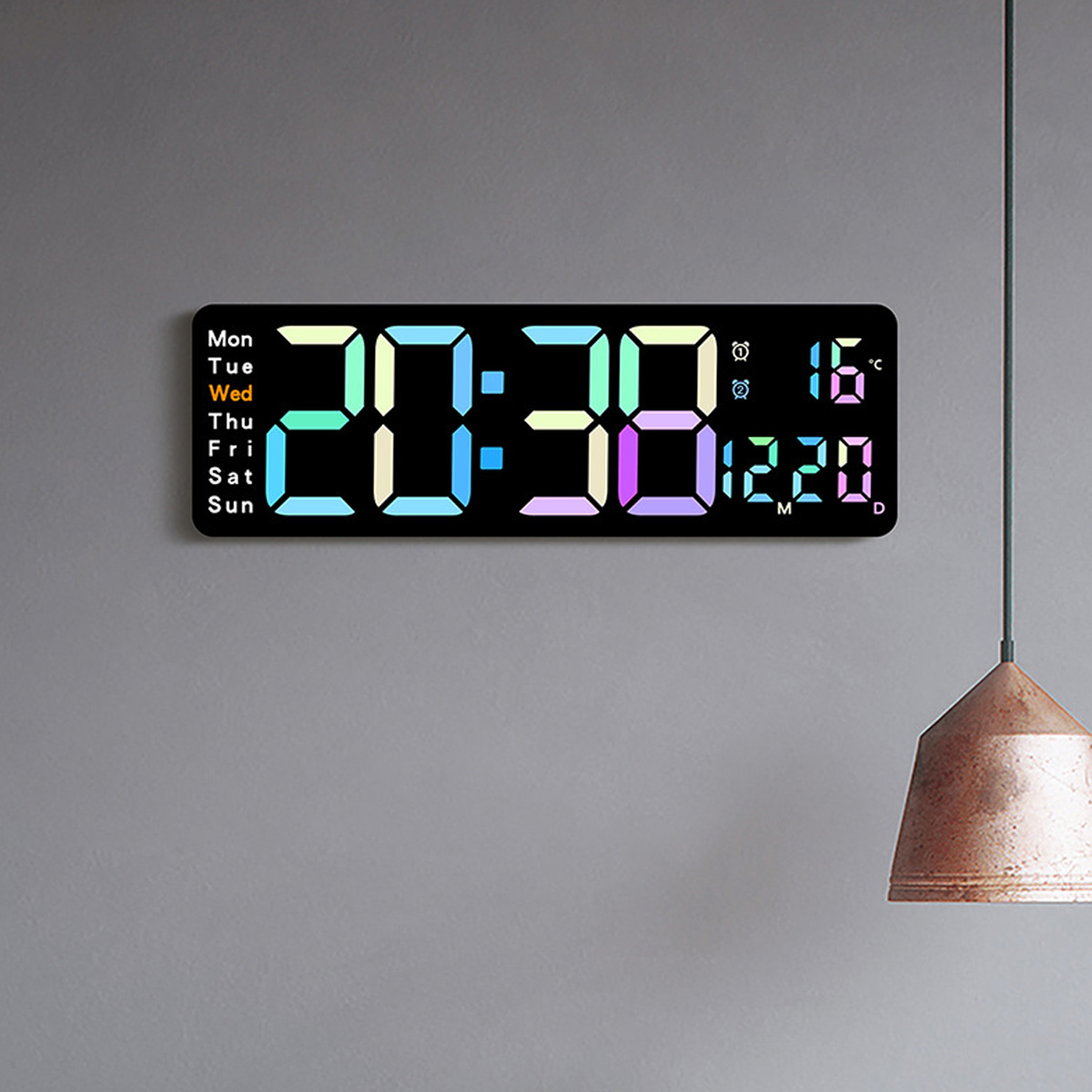 Digitaluhr mit Anzeige von Uhrzeit, Datum und Temperatur. Die Uhr befindet sich an einer grauen Wand. Eine Hängelampe ist in der rechten Ecke.